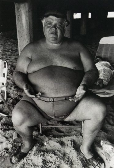 A shirtless man with a prominent belly and a cap sits on a beach, looking directly at the viewer. His hands rest on his swimming trunks, and a column is visible behind him.