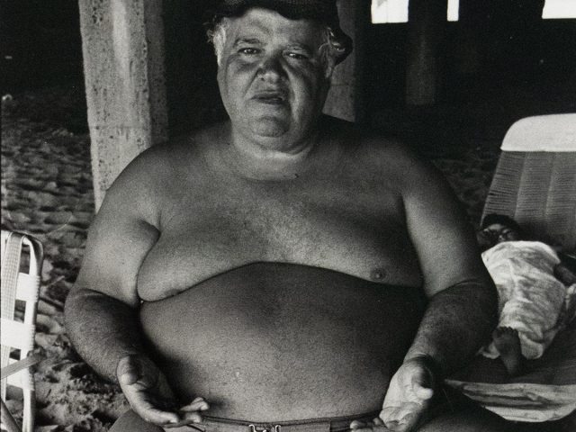 A shirtless man with a prominent belly and a cap sits on a beach, looking directly at the viewer. His hands rest on his swimming trunks, and a column is visible behind him.