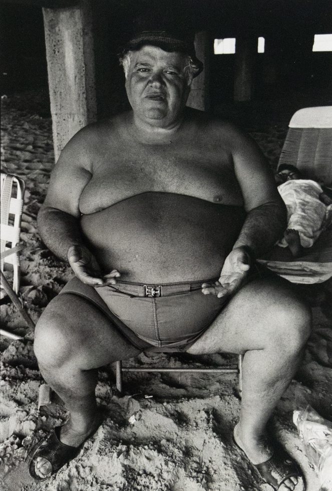 A shirtless man with a prominent belly and a cap sits on a beach, looking directly at the viewer. His hands rest on his swimming trunks, and a column is visible behind him.