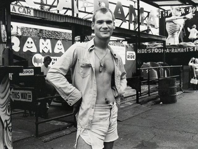 A black and white photograph shows a young man with a slight smile standing in front of an amusement park ride entrance. He wears an open collared shirt, shorts, and a necklace.