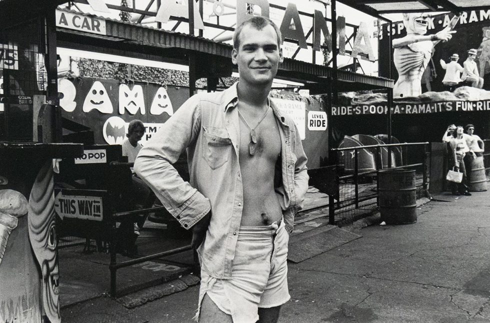 A black and white photograph shows a young man with a slight smile standing in front of an amusement park ride entrance. He wears an open collared shirt, shorts, and a necklace.