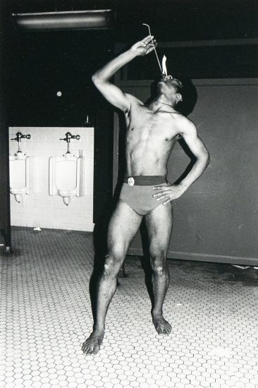 A black and white photograph captures a shirtless man in a small bathing suit in a men's restroom, holding a flaming stick above his mouth. Two urinals are visible on the left side of the frame.