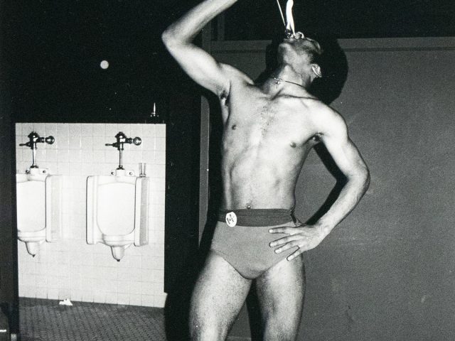 A black and white photograph captures a shirtless man in a small bathing suit in a men's restroom, holding a flaming stick above his mouth. Two urinals are visible on the left side of the frame.