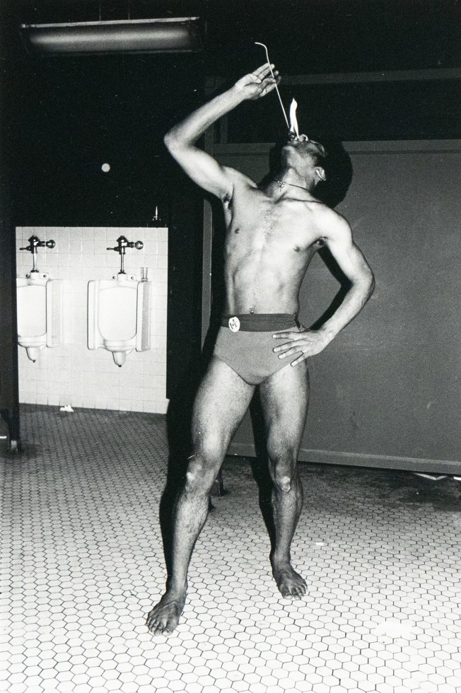 A black and white photograph captures a shirtless man in a small bathing suit in a men's restroom, holding a flaming stick above his mouth. Two urinals are visible on the left side of the frame.