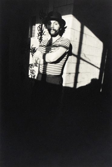 A black and white photograph captures a man with a beard and hat, wearing a striped shirt and dark pants, standing with his arms crossed in a strong shadow. Graffiti is visible on the wall behind him.