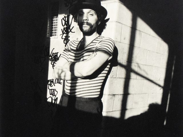 A black and white photograph captures a man with a beard and hat, wearing a striped shirt and dark pants, standing with his arms crossed in a strong shadow. Graffiti is visible on the wall behind him.