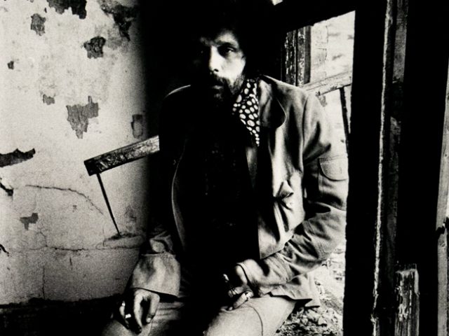 A black and white photograph captures a person with an Afro and beard, wearing a jacket and polka-dotted scarf, sitting in a dilapidated window frame. The light illuminates their face and the textured, crumbling wall behind them.