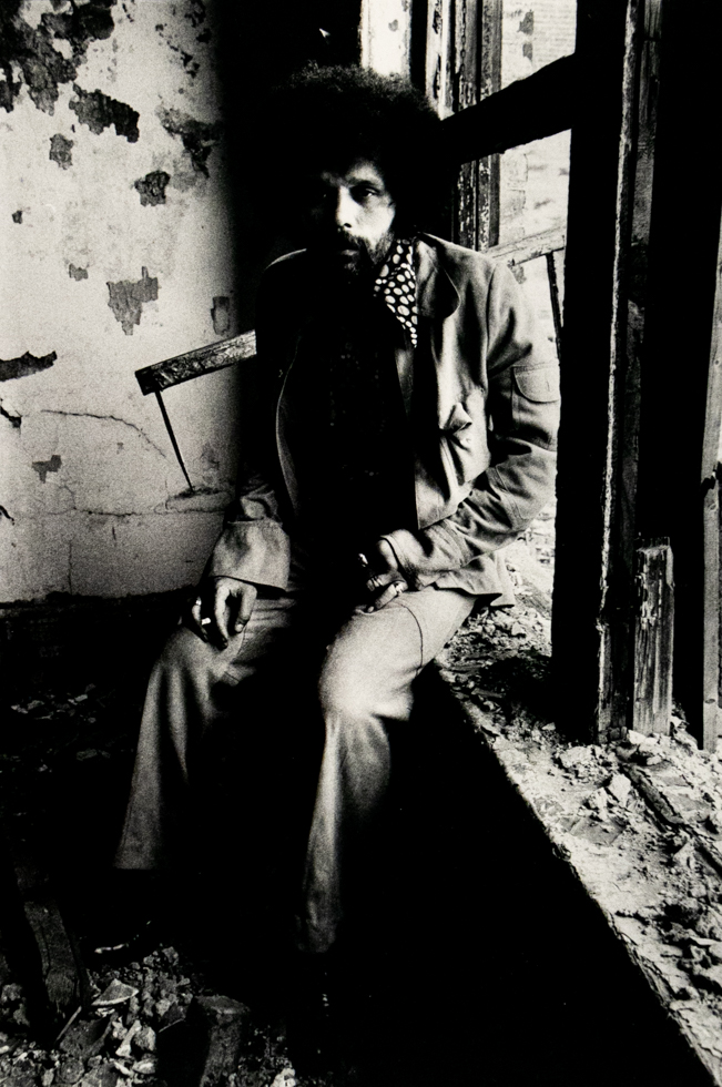 A black and white photograph captures a person with an Afro and beard, wearing a jacket and polka-dotted scarf, sitting in a dilapidated window frame. The light illuminates their face and the textured, crumbling wall behind them.