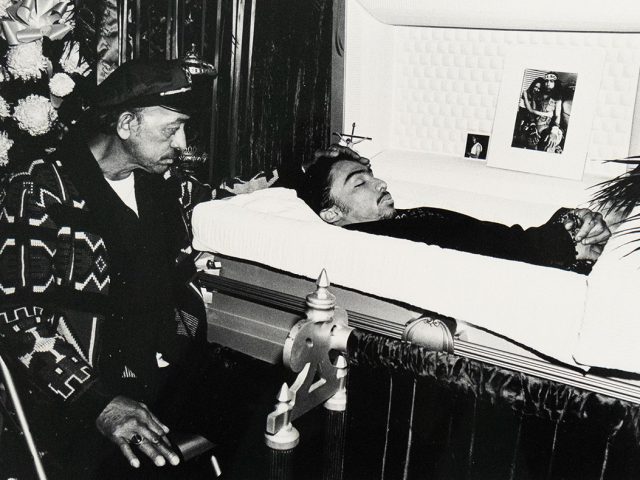 A man wearing a cap and patterned jacket sits beside an open casket, looking at the deceased within. The deceased is shown from the chest up, and photographs are displayed near the head of the casket.