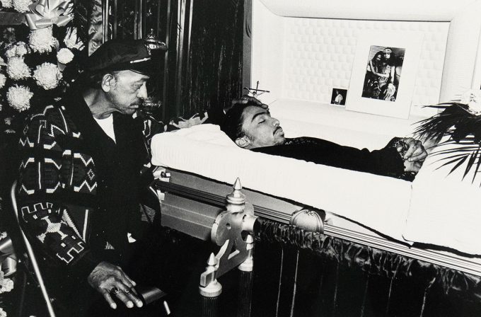 A man wearing a cap and patterned jacket sits beside an open casket, looking at the deceased within. The deceased is shown from the chest up, and photographs are displayed near the head of the casket.