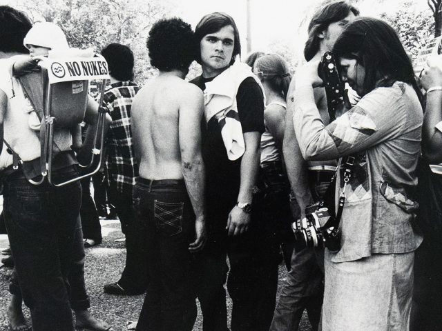 A black and white photograph captures a crowd of people outdoors. In the foreground, a person on the left carries a child in a backpack with a "NO NUKES" sign, while other individuals, some with cameras, are visible in the busy scene.