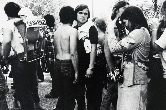 A black and white photograph captures a crowd of people outdoors. In the foreground, a person on the left carries a child in a backpack with a "NO NUKES" sign, while other individuals, some with cameras, are visible in the busy scene.