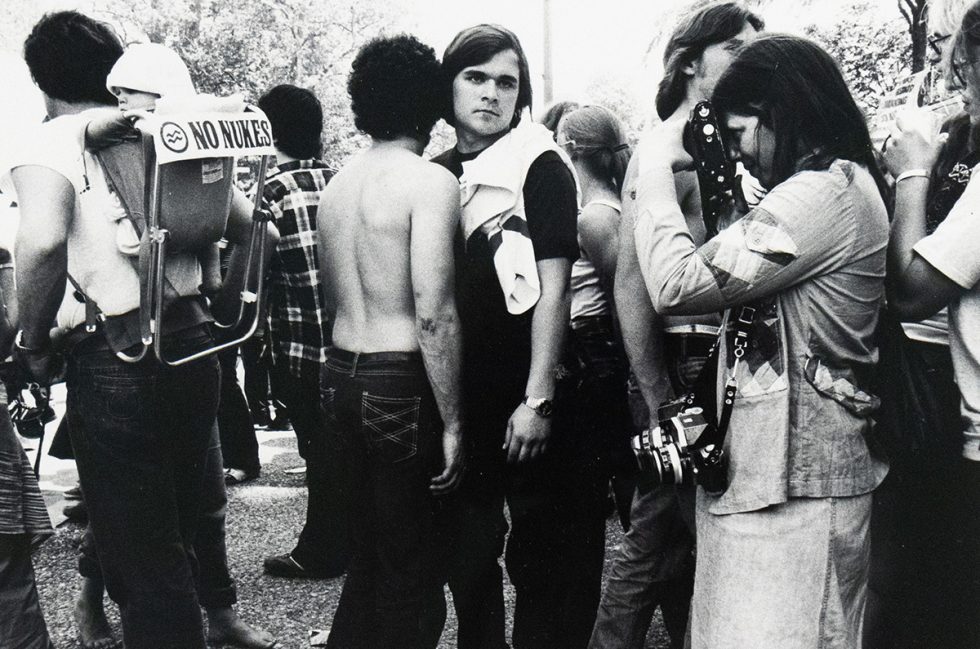 A black and white photograph captures a crowd of people outdoors. In the foreground, a person on the left carries a child in a backpack with a "NO NUKES" sign, while other individuals, some with cameras, are visible in the busy scene.