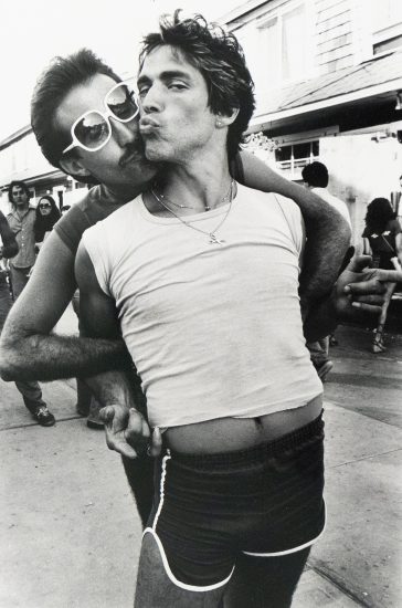 A black and white photograph captures two men embracing on a street. One man, wearing sunglasses, kisses the other man's cheek, who puckers his lips toward the viewer.