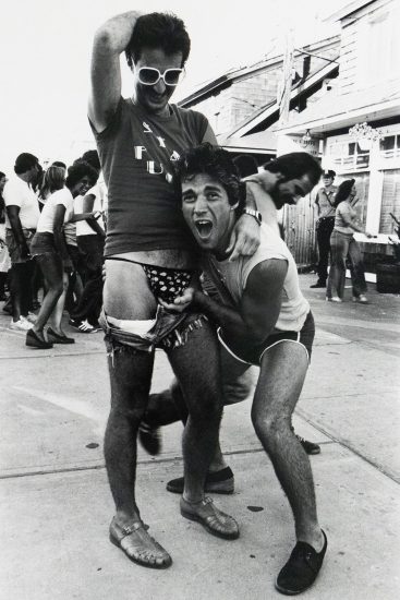 A black and white photograph of two men playfully posing on a boardwalk, with one lifting the other's shorts to reveal patterned underwear as they smile and interact with the camera. People in casual summer clothing walk and laugh in the background.