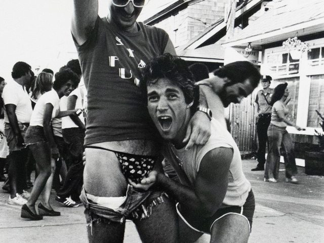 A black and white photograph of two men playfully posing on a boardwalk, with one lifting the other's shorts to reveal patterned underwear as they smile and interact with the camera. People in casual summer clothing walk and laugh in the background.
