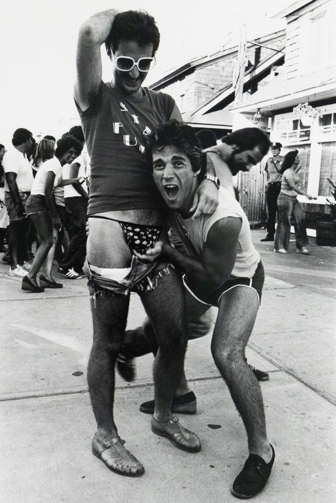 A black and white photograph of two men playfully posing on a boardwalk, with one lifting the other's shorts to reveal patterned underwear as they smile and interact with the camera. People in casual summer clothing walk and laugh in the background.