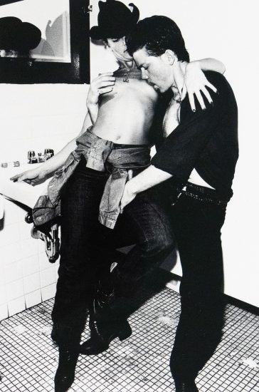 A shirtless woman in a cowboy hat leans against a tiled bathroom sink as a man holds her leg and presses in toward her. A small mirror and sink fixtures are visible in the background.
