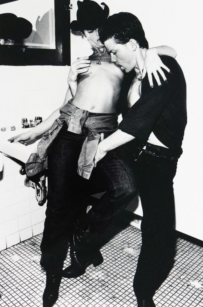 A shirtless woman in a cowboy hat leans against a tiled bathroom sink as a man holds her leg and presses in toward her. A small mirror and sink fixtures are visible in the background.