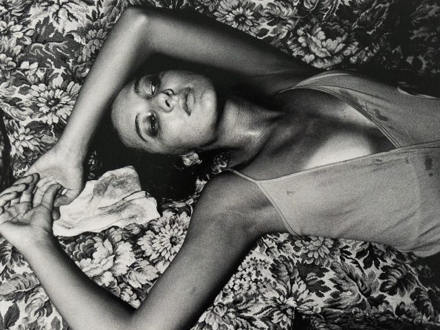 Black-and-white photograph of a woman lying on a patterned floral couch with her arms raised above her head. She wears a sleeveless garment and gazes upward with a cloth beneath one hand.