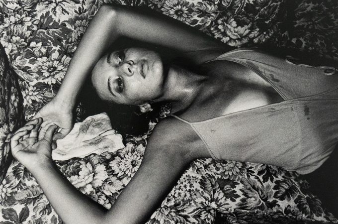 Black-and-white photograph of a woman lying on a patterned floral couch with her arms raised above her head. She wears a sleeveless garment and gazes upward with a cloth beneath one hand.