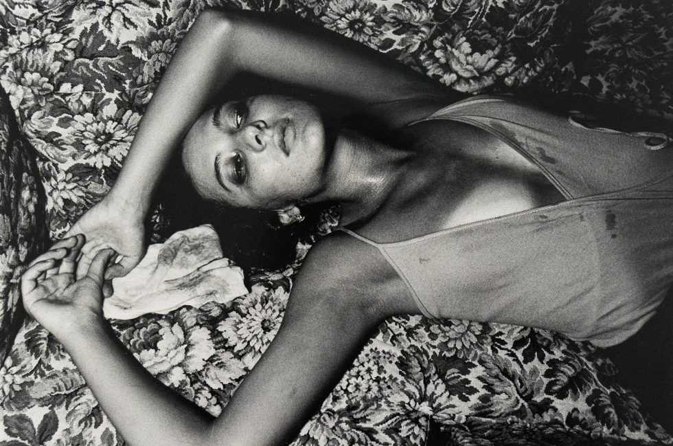 Black-and-white photograph of a woman lying on a patterned floral couch with her arms raised above her head. She wears a sleeveless garment and gazes upward with a cloth beneath one hand.