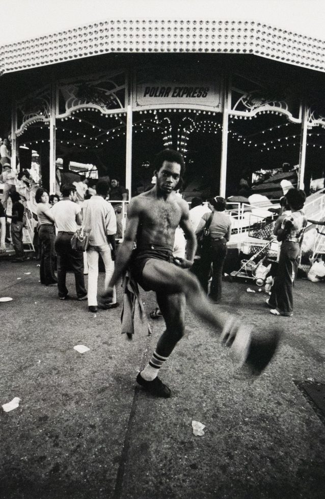 A black and white photograph captures a shirtless man mid-stride with one leg extended, looking at the viewer. He is in front of a carnival ride named "Polar Express," with other people and debris visible on the ground.