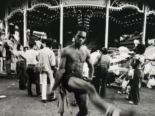 A black and white photograph captures a shirtless man mid-stride with one leg extended, looking at the viewer. He is in front of a carnival ride named "Polar Express," with other people and debris visible on the ground.