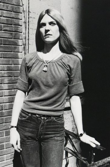 A black and white photograph captures a woman in a casual outfit, standing against a brick wall with her left hand resting on a metal railing. She wears a gathered-neck shirt, jeans, and a necklace, looking directly forward with a somber expression.