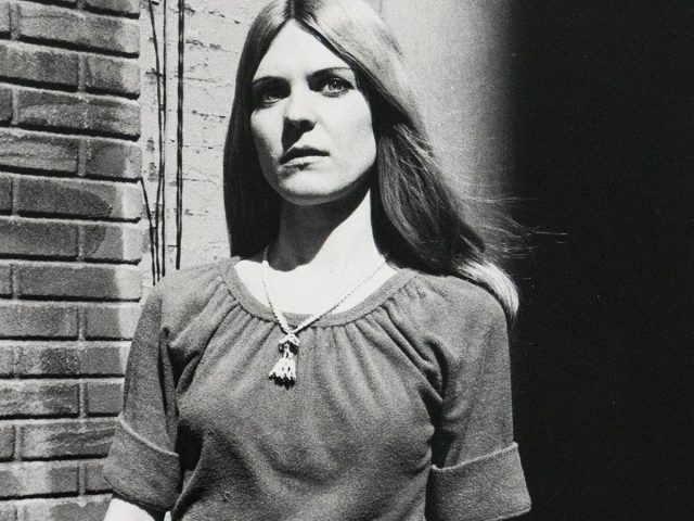 A black and white photograph captures a woman in a casual outfit, standing against a brick wall with her left hand resting on a metal railing. She wears a gathered-neck shirt, jeans, and a necklace, looking directly forward with a somber expression.
