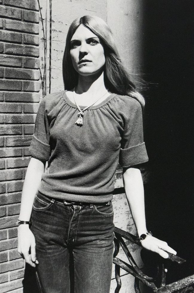 A black and white photograph captures a woman in a casual outfit, standing against a brick wall with her left hand resting on a metal railing. She wears a gathered-neck shirt, jeans, and a necklace, looking directly forward with a somber expression.