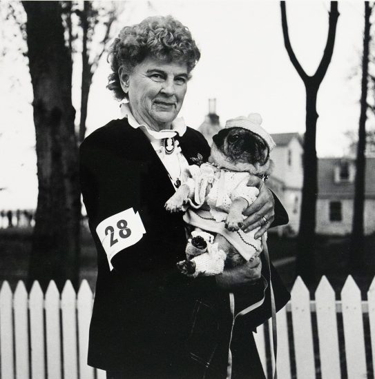 A black and white photograph captures an older woman with curly hair, wearing a dark jacket with a "28" bib, holding a pug dressed in a knitted outfit and hat. They are outdoors, with a white picket fence, trees, and a house in the background.