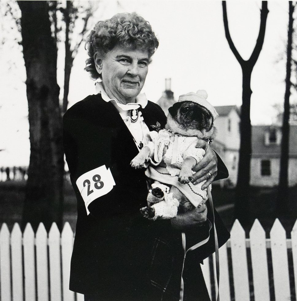 A black and white photograph captures an older woman with curly hair, wearing a dark jacket with a "28" bib, holding a pug dressed in a knitted outfit and hat. They are outdoors, with a white picket fence, trees, and a house in the background.
