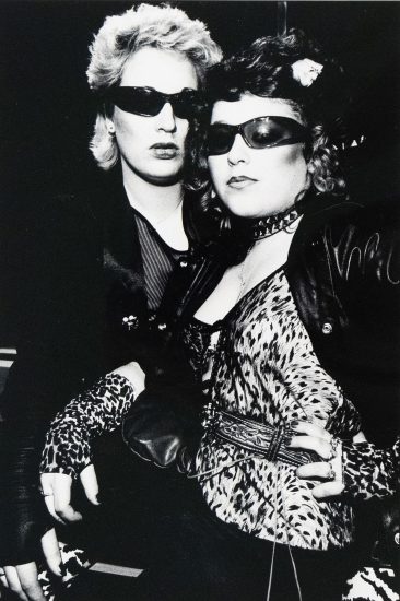 A black and white photograph captures two individuals, both wearing sunglasses, posing closely together. The person in the foreground wears a leopard print top and gloves, adorned with a studded choker and a patterned belt.
