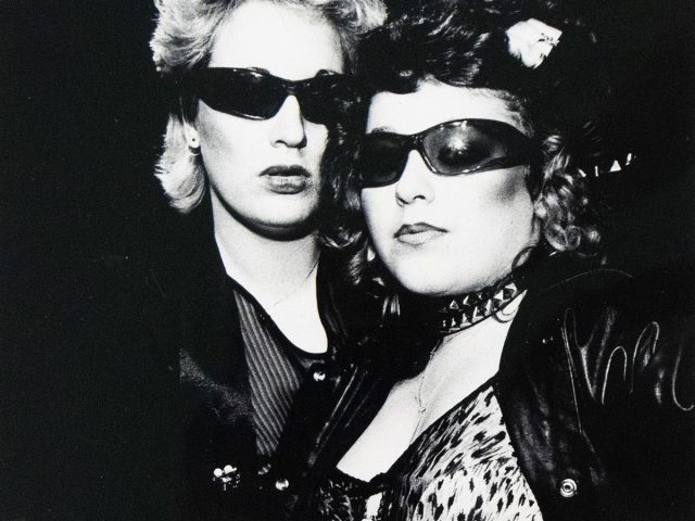 A black and white photograph captures two individuals, both wearing sunglasses, posing closely together. The person in the foreground wears a leopard print top and gloves, adorned with a studded choker and a patterned belt.
