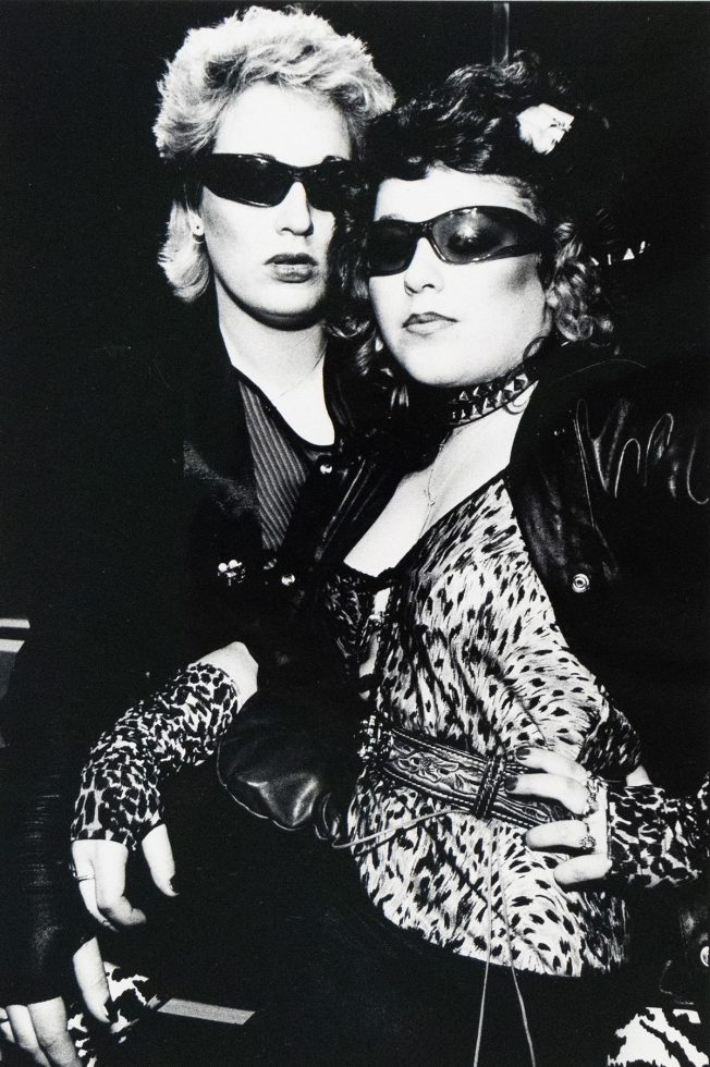 A black and white photograph captures two individuals, both wearing sunglasses, posing closely together. The person in the foreground wears a leopard print top and gloves, adorned with a studded choker and a patterned belt.