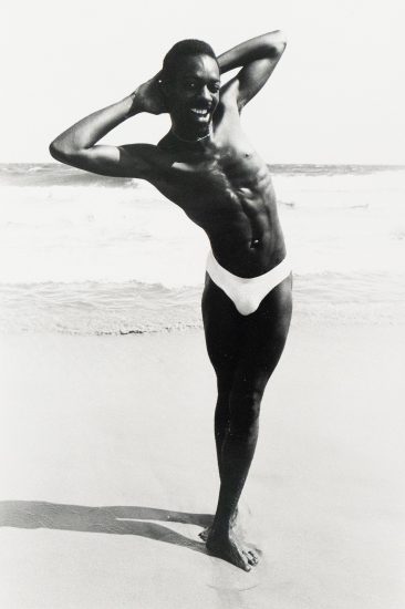 A black and white photograph captures a shirtless man in a speedo posing on a beach with his arms behind his head. He smiles broadly, with the ocean and sky visible in the background.