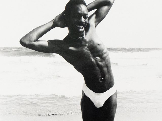 A black and white photograph captures a shirtless man in a speedo posing on a beach with his arms behind his head. He smiles broadly, with the ocean and sky visible in the background.