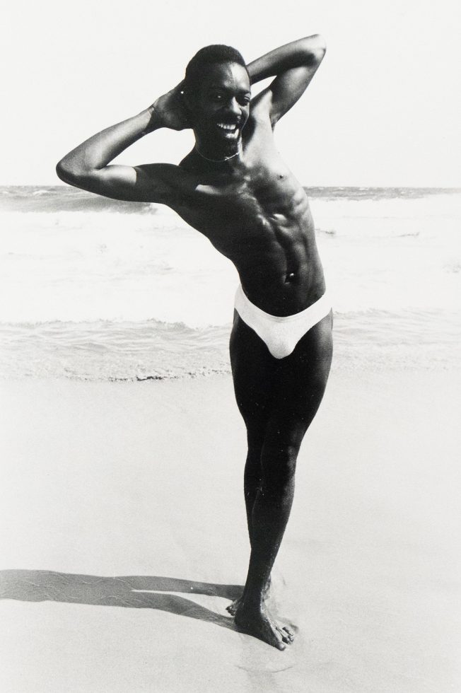 A black and white photograph captures a shirtless man in a speedo posing on a beach with his arms behind his head. He smiles broadly, with the ocean and sky visible in the background.