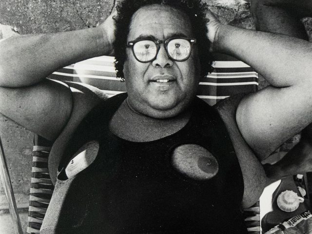 A person with curly hair and glasses lies on a striped beach chair, seen from a high angle. They are wearing a dark, one-piece swimsuit with circular cutouts at the chest, exposing the nipples.