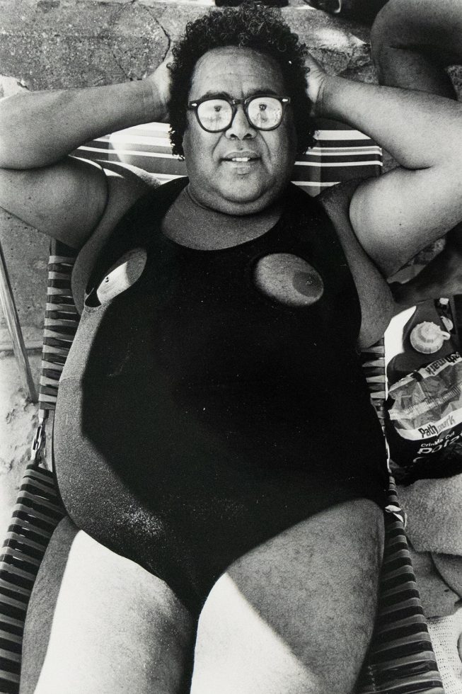A person with curly hair and glasses lies on a striped beach chair, seen from a high angle. They are wearing a dark, one-piece swimsuit with circular cutouts at the chest, exposing the nipples.