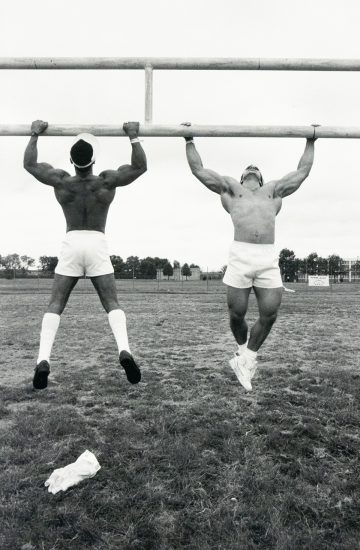 A black and white photograph captures two muscular, shirtless men hanging from a horizontal bar, mid-air, on a grassy field. The man on the left faces away, while the man on the right faces towards the camera.