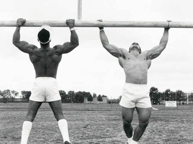 A black and white photograph captures two muscular, shirtless men hanging from a horizontal bar, mid-air, on a grassy field. The man on the left faces away, while the man on the right faces towards the camera.
