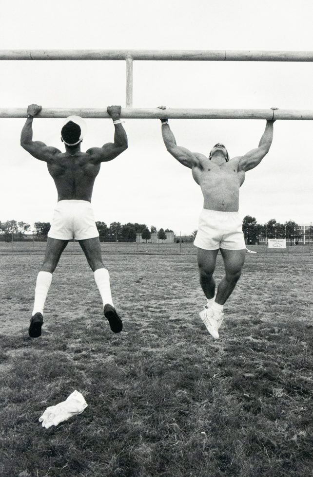 A black and white photograph captures two muscular, shirtless men hanging from a horizontal bar, mid-air, on a grassy field. The man on the left faces away, while the man on the right faces towards the camera.