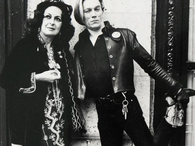 A black and white photograph captures two individuals standing against a textured wall. A woman in a dark, embroidered dress and hat stands next to a man with light hair, who wears a leather jacket and one boot raised.