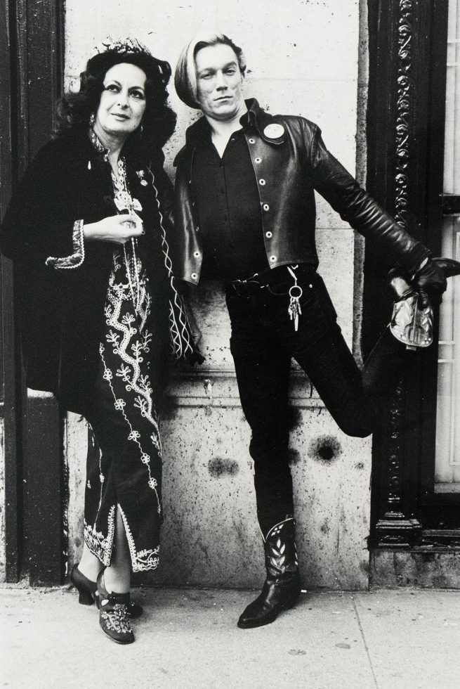 A black and white photograph captures two individuals standing against a textured wall. A woman in a dark, embroidered dress and hat stands next to a man with light hair, who wears a leather jacket and one boot raised.