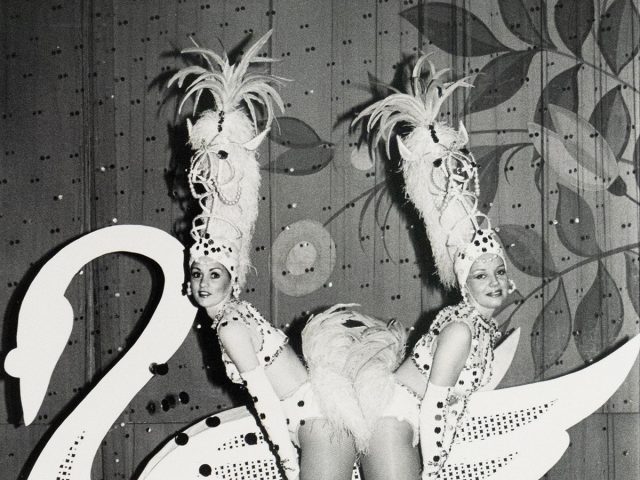 A duo of performers in feathered costumes and headpieces pose behind a large swan prop on a stage. They are looking over their shoulders at the viewer.