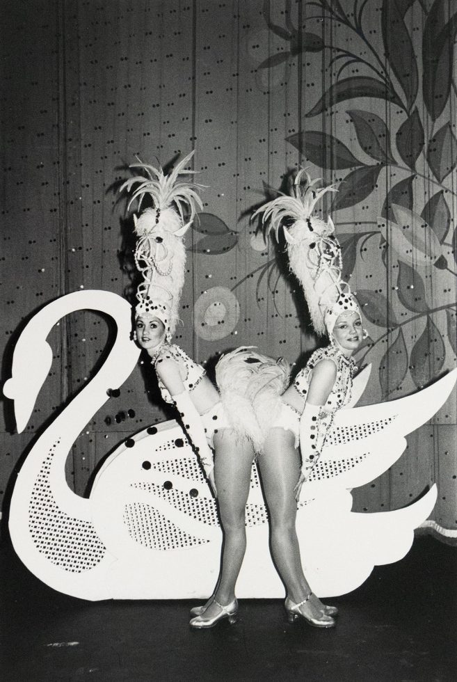 A duo of performers in feathered costumes and headpieces pose behind a large swan prop on a stage. They are looking over their shoulders at the viewer.
