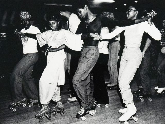 A group of roller skaters, predominantly Black, are captured in a black and white photograph performing a line dance at an indoor rink. They are holding hands and extending their arms, moving in unison across the frame.