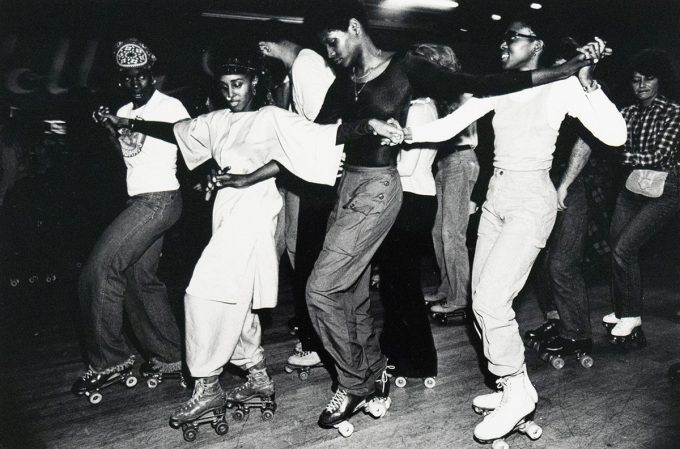 A group of roller skaters, predominantly Black, are captured in a black and white photograph performing a line dance at an indoor rink. They are holding hands and extending their arms, moving in unison across the frame.